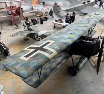 A camouflaged WW1 biplane in a hanger with other aircraft behind