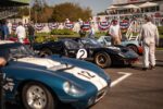 Three race cars at the start line for the 2023 Goodwood Revival, with spectators in the background and race team members standing around on the tarmac. The centre car is a sleek black Ford GT MkII