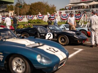 Three race cars at the start line for the 2023 Goodwood Revival, with spectators in the background and race team members standing around on the tarmac. The centre car is a sleek black Ford GT MkII