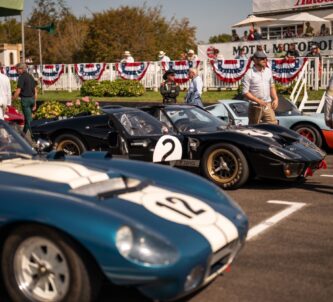 Three race cars at the start line for the 2023 Goodwood Revival, with spectators in the background and race team members standing around on the tarmac. The centre car is a sleek black Ford GT MkII