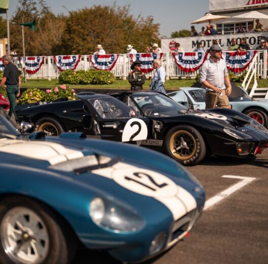Three race cars at the start line for the 2023 Goodwood Revival, with spectators in the background and race team members standing around on the tarmac. The centre car is a sleek black Ford GT MkII