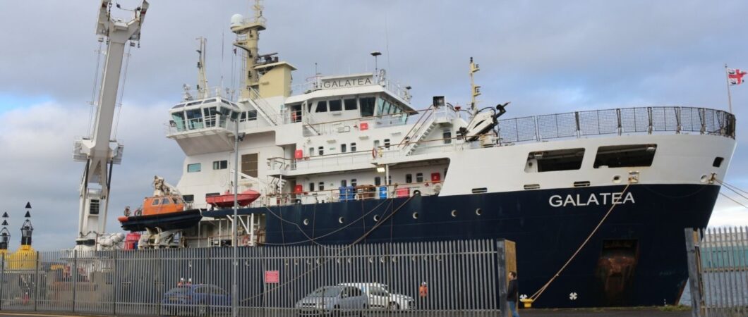 THV Galatea tied up alongside a quay. She's one of those modern ships with a high bow and superstructure at the front. The hull is dark blue and her superstructure is white. There are some cars and yellow navigation buoys (cardinal marks) on the quayside