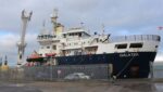 THV Galatea tied up alongside a quay. She's one of those modern ships with a high bow and superstructure at the front. The hull is dark blue and her superstructure is white. There are some cars and yellow navigation buoys (cardinal marks) on the quayside