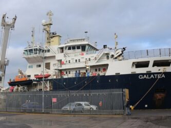 THV Galatea tied up alongside a quay. She's one of those modern ships with a high bow and superstructure at the front. The hull is dark blue and her superstructure is white. There are some cars and yellow navigation buoys (cardinal marks) on the quayside