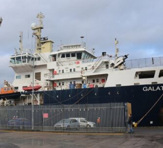 THV Galatea tied up alongside a quay. She's one of those modern ships with a high bow and superstructure at the front. The hull is dark blue and her superstructure is white. There are some cars and yellow navigation buoys (cardinal marks) on the quayside