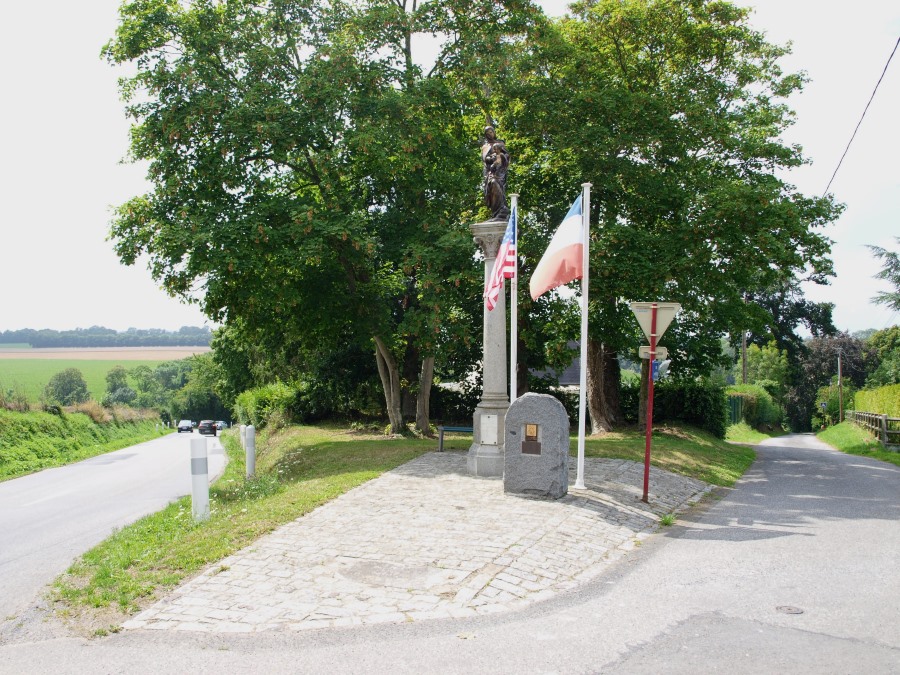 A roadside memorial with a plaque on a stone, an etched copper panel and flagpoles. It is at a junction with roads passing on both sides.
