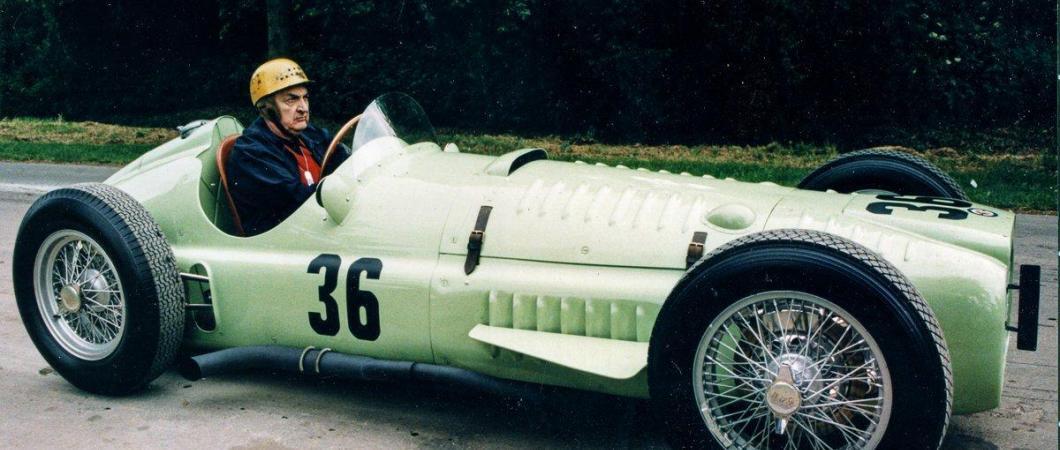 Driver with leather cap sits in a light green 1950s BRM Formula 1 race car