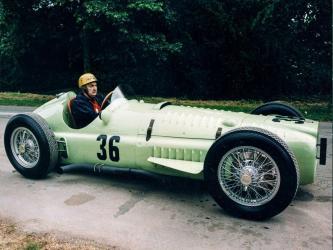 Driver with leather cap sits in a light green 1950s BRM Formula 1 race car