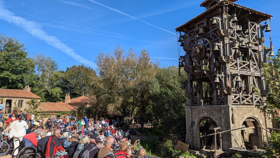 An audience sits in front of a bell tower with exposed workings, waiting for the performance to begin