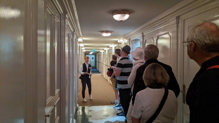 A guide talks to visitors in a ship's corridor of cabin doors