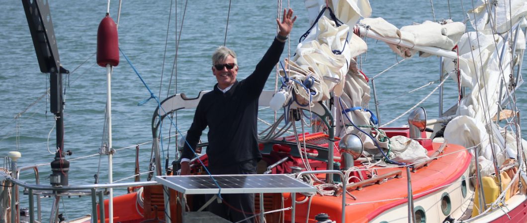 A middle aged yachtsman waves at the camera from the cockpit of his small racing yacht
