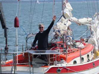 A middle aged yachtsman waves at the camera from the cockpit of his small racing yacht