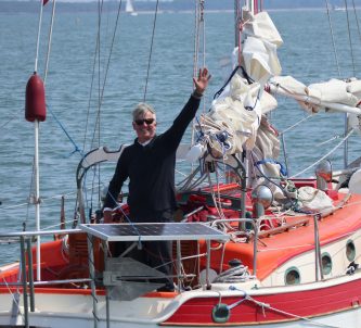 A middle aged yachtsman waves at the camera from the cockpit of his small racing yacht