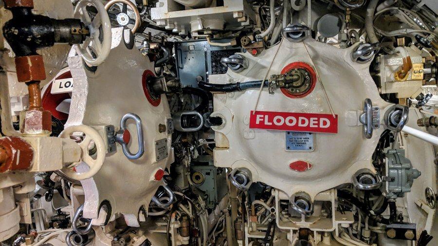Two white-painted torpedo tubes. One is open. The hatch of the other is closed, locked and has a red sign hanging on it "Flooded"