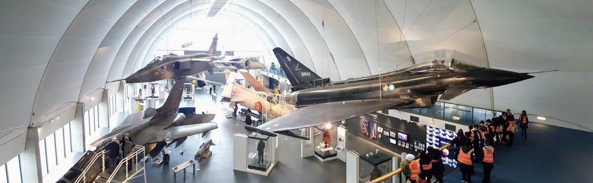 View looking down on large jet aircraft exhibited on the floor and suspended. There's a group of school children in hi-viz jackets looking on from a lower balcony.