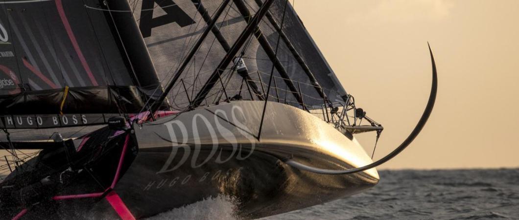 A black Vendee Globe racing yacht, Hugo Boss, heels over exposing her foils in the evening sunlight