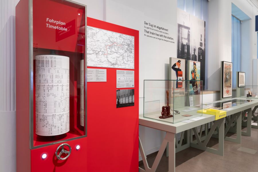 A large red cabinet has a railway timetable printed on a tall cylinder with a handwheel to rotate it. Behind are other objects on display.