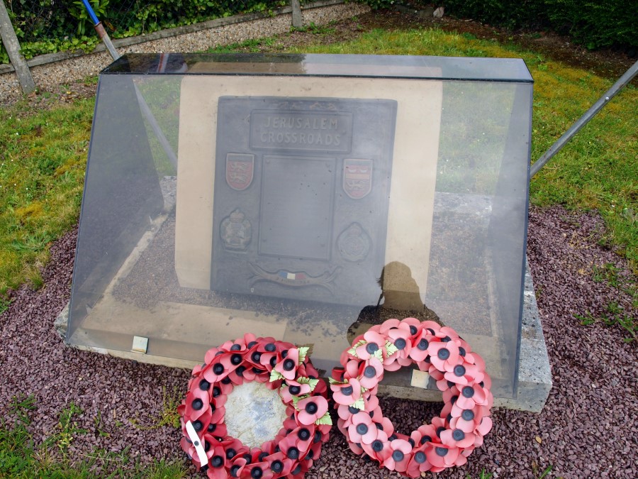 A small stone with a bronze plaque covered with a dark perspex container. There are wreaths propped up against it.