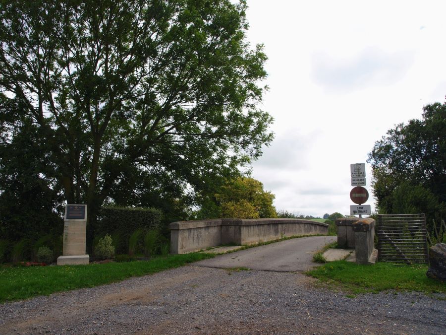 View of the small road bridge and the memorial marker stone beside it, under the shade of a large tree