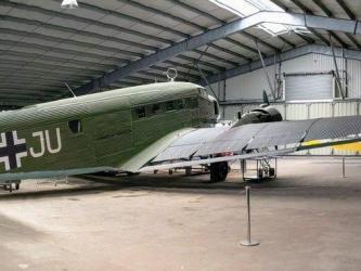 A three engined Junkers 52 transport aircraft in Luftwaffe colours, dark green and yellow, parked in a large hanger at the Salis Flying Museum
