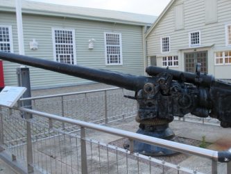 A black-painted naval gun mounted on a plinth in front of some wooden museum buildings