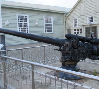 A black-painted naval gun mounted on a plinth in front of some wooden museum buildings