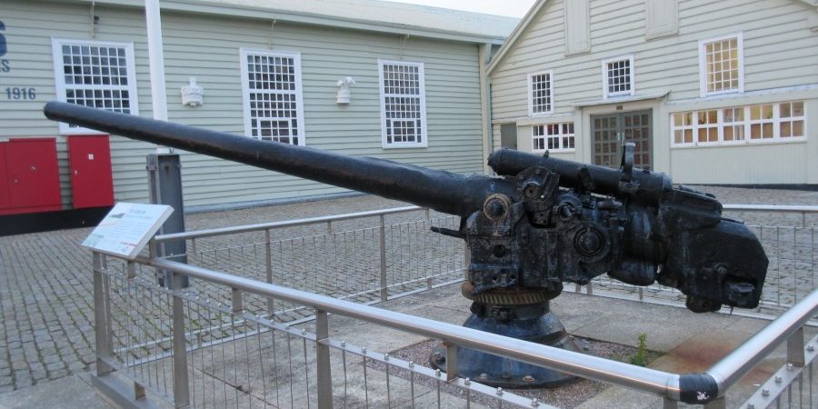 A black-painted naval gun mounted on a plinth in front of some wooden museum buildings