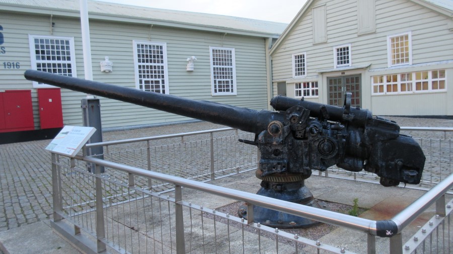 A black-painted naval gun mounted on a plinth in front of some wooden museum buildings