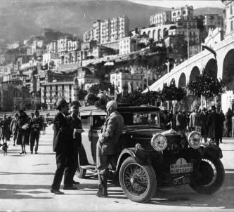 A black & white photo of Kitty Brunell's Talbot car parked on the quayside at Monte Carlo. She is leaning over the open roof talking to three men in suits.