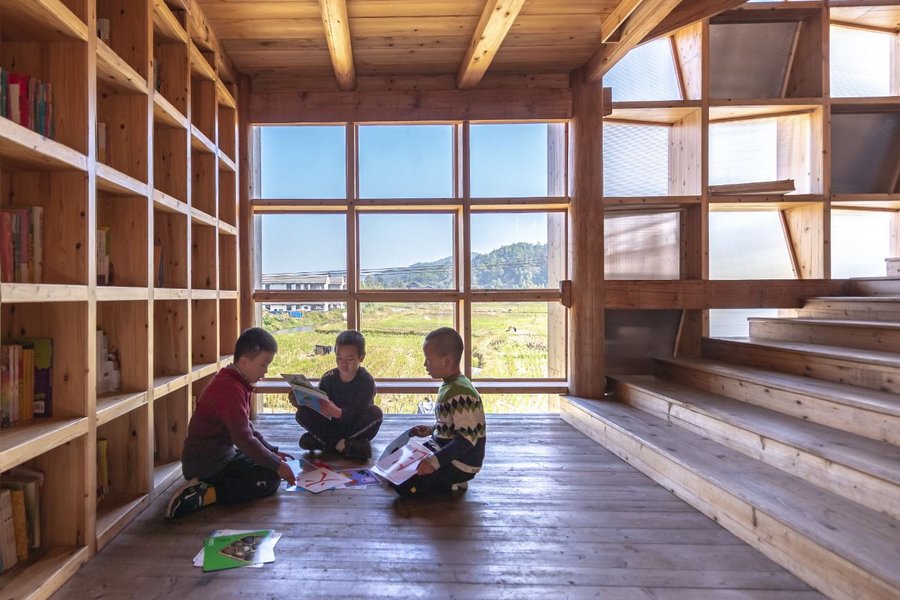Children sit on a wooden floor beneath bookshelves. Behind them is a window looking out onto rice paddy fields and mountains.