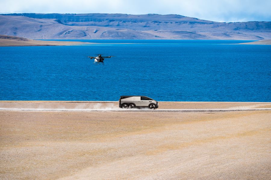 A silver 6-wheel vehicle speeds along a dusty desert road beside the sea shore with a small multi-rotor eVTOL in the air behind it.