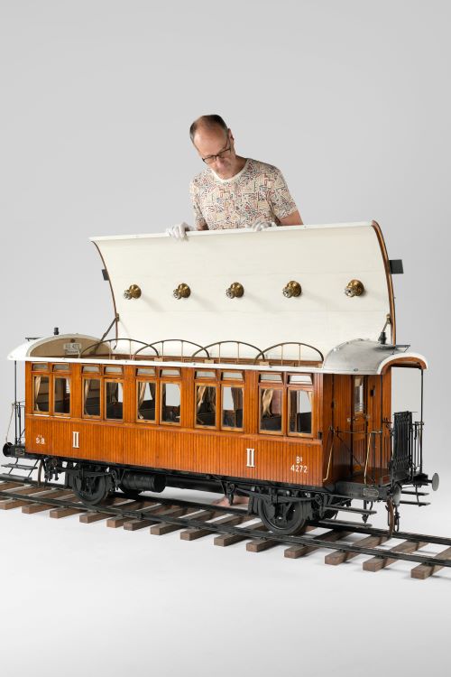 A man holds open the rook of a model late 19th century railway carriage, which would be significantly longer than him if he laid down beside it.