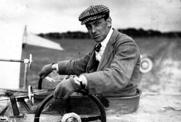 Black and white close-up of Latham in jacket, tie and with a cap, in his aircraft