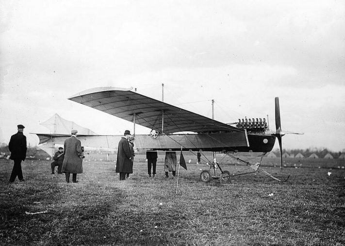 Black & white photo of men in coats, caps & hats, standing around a monoplane in a field