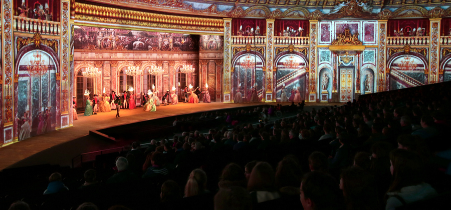 The audience in the foreground watch a scene with a large 18th century palace boardroom with lords & ladies dancing. The ladies have illuminated ships on their heads!