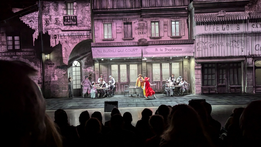 The two main characters in red and yellow costumes dance at a street cafe with waiters and customers