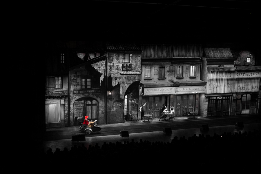 The two main characters in red and yellow costumes and riding a motorcycle and sidecar drive through black & white streets