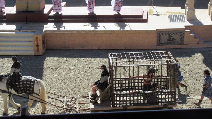 A prisoner in a cage on a cart is paraded, with two others attached to the back of the cart by chains