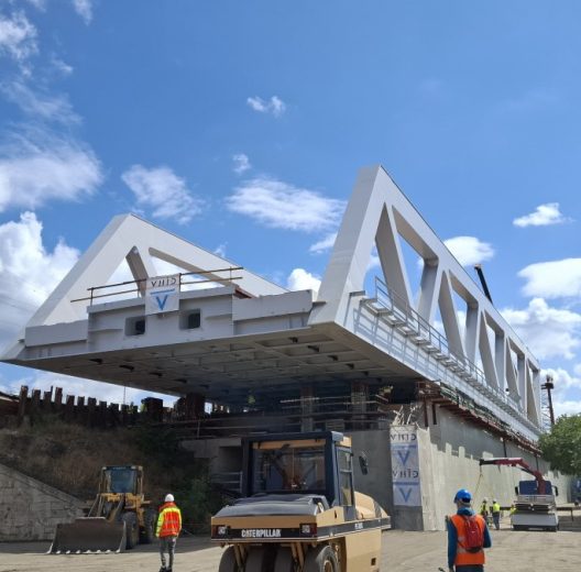 A grey steel box girder bridge span hangs over the road