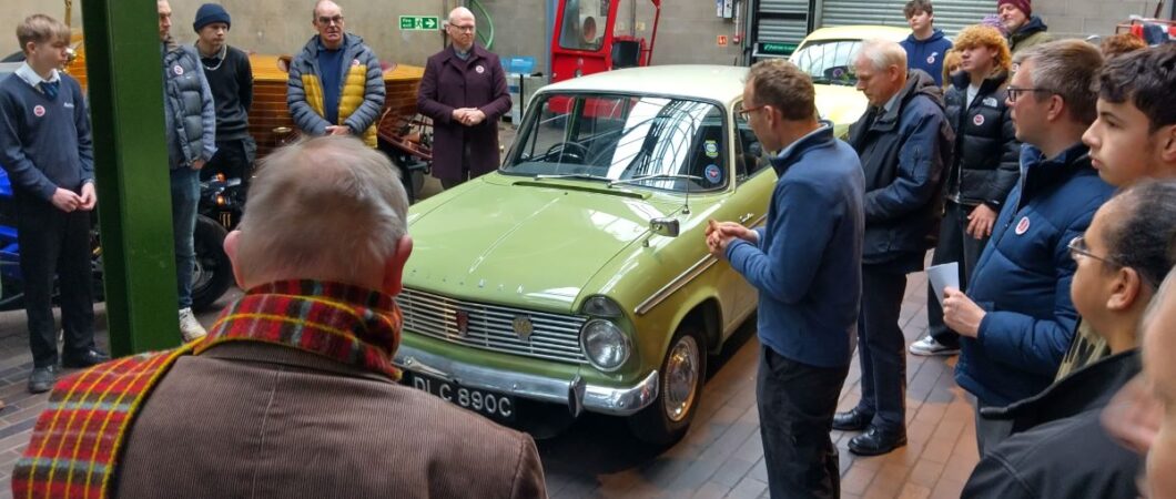 A green Hillman car surrounded by people in a garage