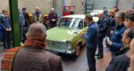 A green Hillman car surrounded by people in a garage