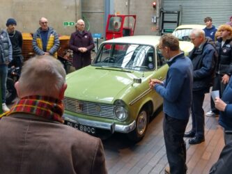 A green Hillman car surrounded by people in a garage