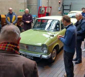 A green Hillman car surrounded by people in a garage