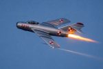A 1950s silver MiG-17 in Polish markings, streaks through the evening sky with a bright flame from the afterburner.