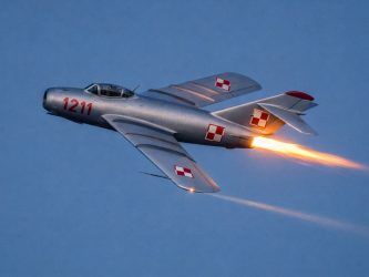 A 1950s silver MiG-17 in Polish markings, streaks through the evening sky with a bright flame from the afterburner.