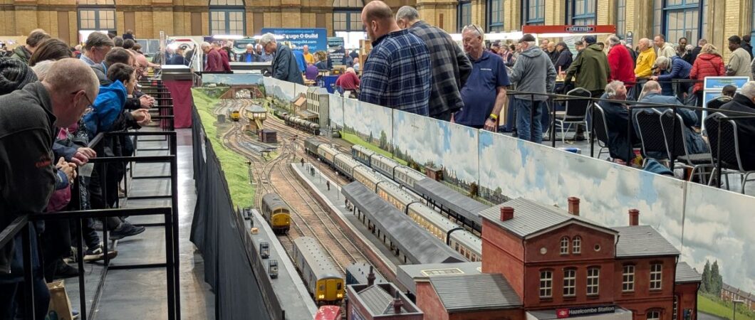 A scale model railway layout at an exhibition, with operators behind it and eager watchers crammed in front