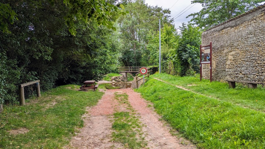 A dirt track leads down to a small river crossing surrounded by trees. There is a picnic table set on grass to the left of the track and a stone building on the right