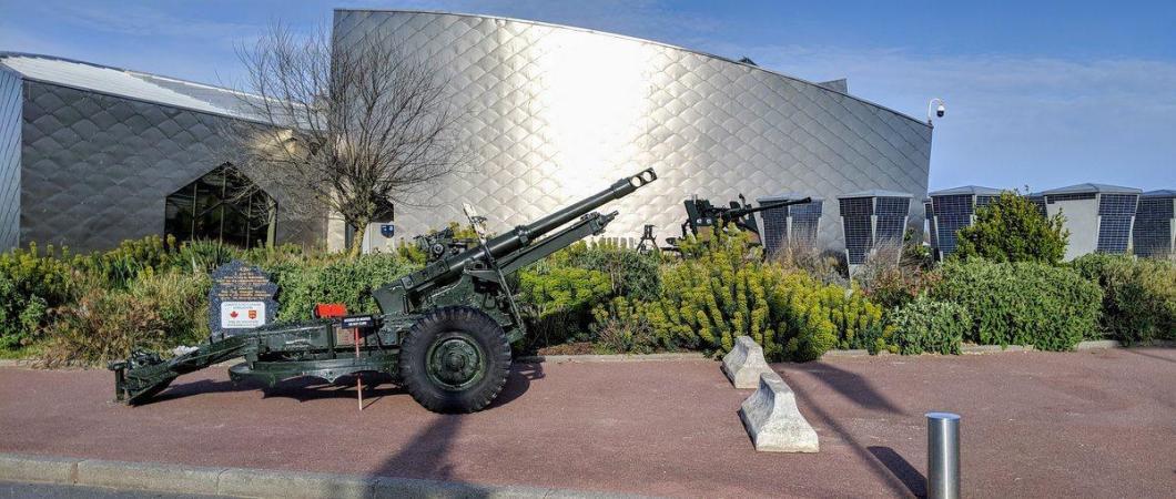 A green field gun sits in front of the curved silver wall of the Juno Beach Center, under a blue sky