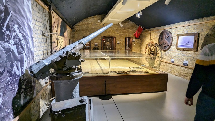A light grey naval gun stands in front of a large model of the evacuation at Dunkirk