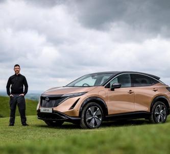 Chris Ramsey stand next to a gold coloured Nissan Ariya in a moorland setting